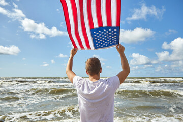 Man in white outfit standing with American flag waving in the wind on the beach.