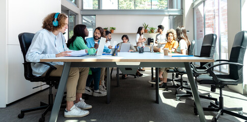 Spacious, luminous, busy office. Multiracial group of coworkers share big table in coworking space.