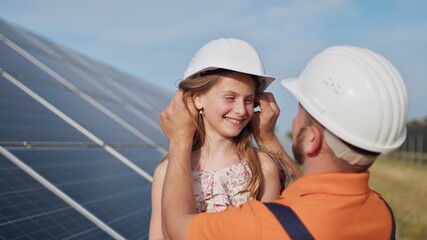 Father with little girl at solar power plant. The father talks about solar energy. The concept of green energy will save the planet for children. The father puts a protective helmet on the girl's head