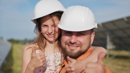 Portrait of the father of a solar power engineer with his little daughter in a protective helmet, pointing thumb up at the camera and smiling. Family at the solar power plant. Planet for children