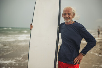 An old man is a surfer with a surfboard. A gray-haired male surfer stands on the beach with a surfboard.