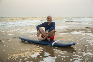 Handsome adult man walks with a surfboard on the seashore. An old surfer with gray hair sits on a surfboard by the sea and looks to the horizon.