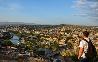 Back view of young man tourist with backpack on the background of panoramic view of Tbilisi city from high point, Georgia
