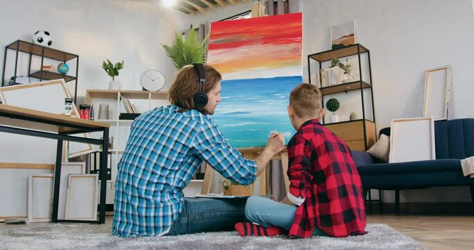Cute boy watching how his talented bearded father -painter drawing picture on canvas in contemporary sitting room