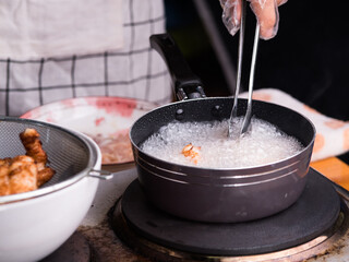 Hands of a woman in gloves are frying the marinated chicken in hot oil in a pan on the kitchen stove and tong the cooked chicken and puts the drained oil on a basket. Selective focus.