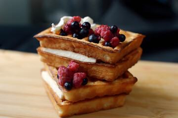 Delicious appetizing waffles with creamy white cream inside lie one on top of the other, covered with ripe raspberries and black currants, just cooked, waiting to be tasted for dessert, close-up photo
