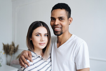 Multiracial couple in love embraces in cozy living room at home. A close-up portrait of smiling caucasian woman and african man are hugging. Love and affection concept