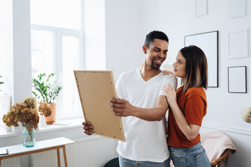 Excited african young husband and caucasian wife hang frame picture on wall happy settling together...