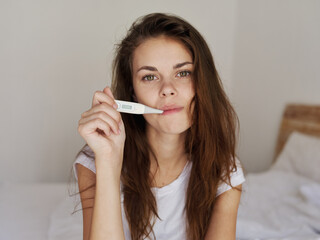woman holding a thermometer in her mouth while sitting on the bed checking temperature