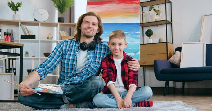 Close up portrait of handsome smiling happy talented bearded man and his son which sitting on the floor near drawn picture on canvas and looking into camera with paintbrushes
