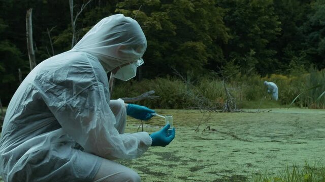 Chemist in ppe looking at flask with solution, water analysis from swamp, wearing protective suit and medical gloves, sample for toxicity testing. Scientific researching, field laboratory concept.
