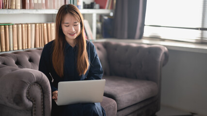 Smiling young woman using laptop,sitting on sofa in living room at home.