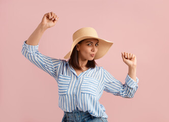 Obraz na płótnie Canvas Lucky pleased woman brunette dances with hands up, has fun, feels freedom and happiness, is champion, won a prize, wears straw hat, striped shirt, jeans, on pink background. Summer emotions concept.