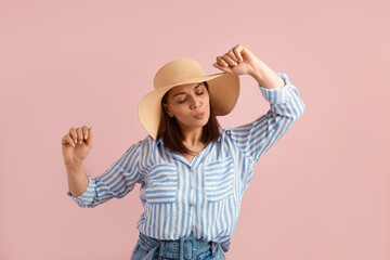 Flirty playful woman brunette in romantic mood dances with hands up, has fun, feels love and happiness, wears straw hat, striped shirt, jeans, on pink background. Summer emotions concept.