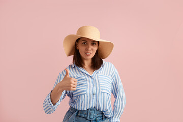 Great job. Satisfied woman cowgirl brunette makes like gesture, keeps thumb up in approval, gives positive feedback, wears straw hat, striped shirt, jeans, on pink background. Summer emotions concept.