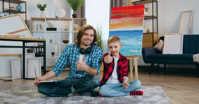 Portrait of handsome smiling happy talented bearded dad and his son both painters which posing on camera in home studio during creating future picture and showing thumbs up