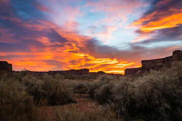 Fototapeta premium The brilliant colors of a sunrise reflecting on clouds over the top of a canyon wall with desert vegetation in the foreground in Utah.