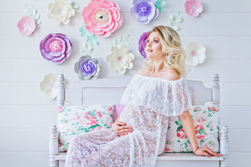 Happy pregnant woman in white interior with decorative flowers. Young blonde woman dreams of a baby. Happy motherhood.