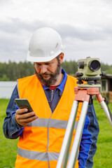 A civil engineer with an optical level looks into a mobile phone. A bearded man is looking for information on his mobile phone.