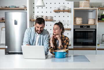 Young smiling man teases his serious girlfriend and shows her a recipe and a photo of what a dish should look like and telling her that she doesn't know how to cook while standing in their kitchen