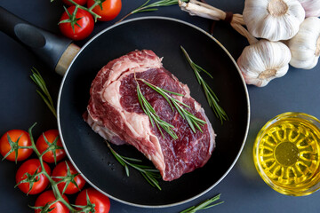 Top view of raw beef meat (Chuck steak ) lying on frying pan on dark table next to red fresh tomatoes, garlic, cooking oil and small green rosemary branches. Selective focus. Fresh organic food theme.