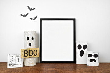 Halloween mock up. Black frame on a wood shelf with rustic wood ghost decor and calendar. Portrait frame against a white wall with bats. Copy space.
