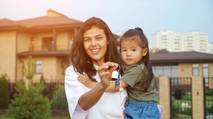 Cheerful woman shows keys of new apartment holding little Korean daughter in arms against detached house in city dwelling district closeup