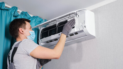 Concentrated electrician in t-shirt and face mask cleans ceiling air conditioner unit with white cloth at maintenance by curtains in room closeup