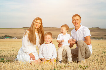 Parents with small sons in white clothes sit on yellow harvested wheat field and rest smiling on hot sunny day against rural landscape, under blue sky