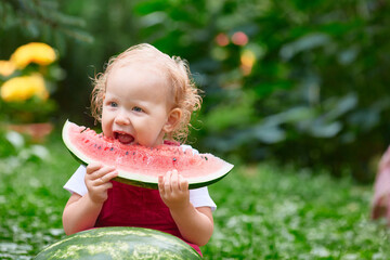 Curly-haired toddler eating watermelon while sitting on green grass in a garden with flowers on a summer day.