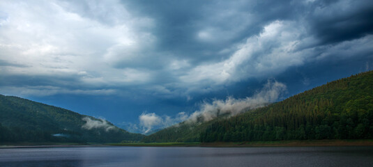 Impressive landscape of green coniferous mountains above lake against the background of stormy gray clouds, rainy weather, Ukraine, Carpathians
