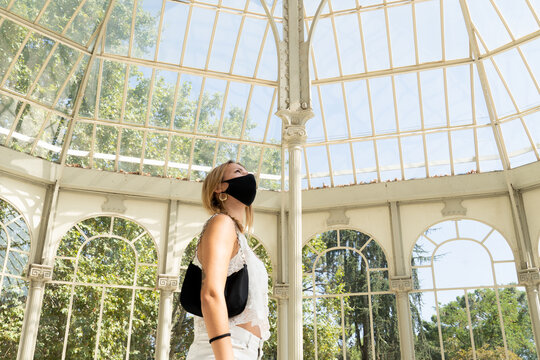 Young blonde white girl dressed in a white tank top with black mask and handbag looking at a glass ceiling