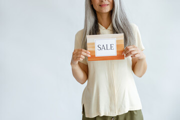 Fototapeta na wymiar Happy middle aged woman with loose silver hair holds colorful Sale sign standing on light grey background in studio closeup, space for text