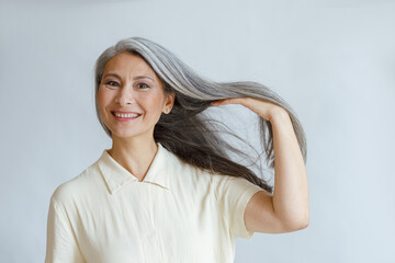 Happy middle aged Asian lady shows natural silver hair posing on light background in studio. Mature...