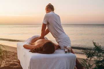 A girl-master of a spa massage at dawn on the seashore makes a back massage to a woman who lies on a massage table. Nearby vases of flowers