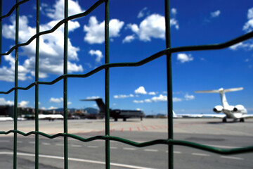 Blurred image of private and business jets parked in the airport apron shot behind a safety net and against a blue sky.