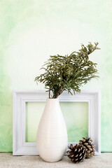 Cedar branch in a white vase against a painterly green background with empty picture frame and pine cones; simple clean design featuring cedar bough and pine cones