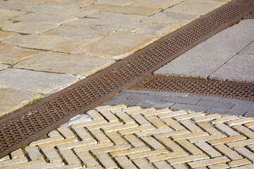 a drainage ditch covered with an iron patterned lattice on a sidewalk made of granite rough tiles...