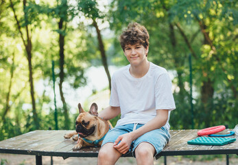Funny french puppy bulldog and teenager playing games outside. Adorable orange bulldog in blue harness in the playground on a sand. Still life, friendship with a dog.