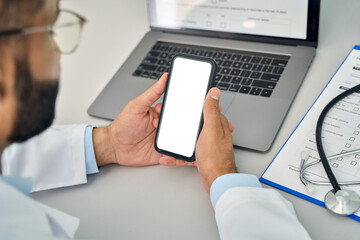 Male doctor gp physician holding mobile cell phone in hands looking at white blank empty mockup smartphone screen working in hospital. Ehealth apps technology mock up concept. Over shoulder view - Powered by Adobe