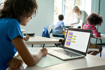 Smart teen kid boy junior middle school student using laptop learning coding data computer science digital software program information technology sitting at desk during class studying in classroom.