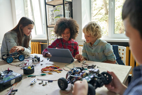 Multiethnic school kids students making robotic cars using tablet computer. Diverse junior children pupils building robot vehicle learning at table at STEM code ai engineering science education class.