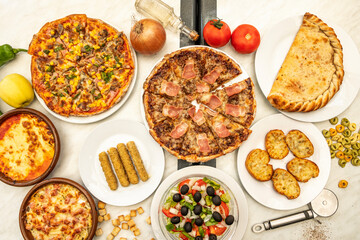 Top view image of Italian food dishes, stuffed calzone, pizza with bacon, garlic bread, Mediterranean salad, mozzarella sticks, tomato and onion, olive oil