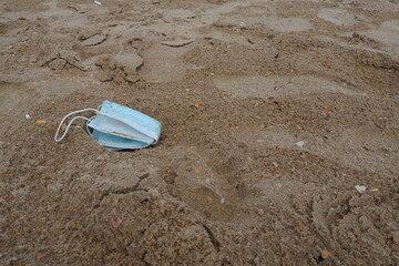 A used medical mask was left on a sandy beach. A dented blue face shield on the sand. Pandemic...