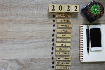 The wooden table on the left has a copy space that represents the timeline of Calendar 2022 and 12 months of wood next to a notebook, pen, smartphone, and cactus.
