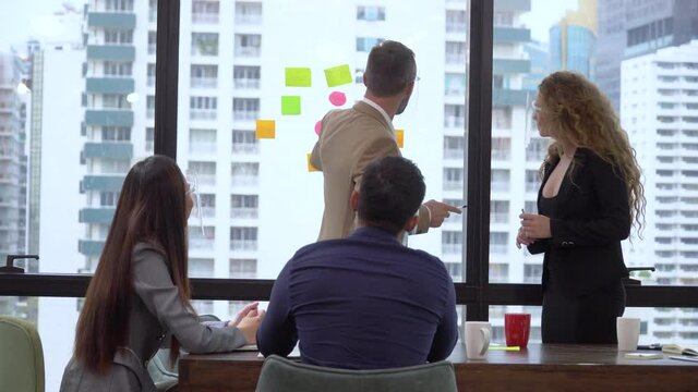 Businessmen And Business Women In Meeting Room Discussing With Information Data With Post It Notes On Window Glass Over Skyscraper Office . Diversity Entrepreneur Talking Strategy On Cityscape View