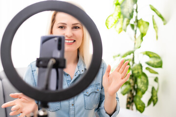 blogger woman recording video on smartphone and lighting herself with ring lamp at table in light...