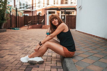 Pretty smiling young woman with tattooes is sitting on the sidewalk. Side view. The concept of relax and psyhology