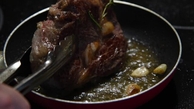Close-up view of chuck steak frying on cooking pan with garlic and rosemary branches. Person fries side of beef steak using metal tongs. Selective focus. 4K resolution video. Food preparation theme.