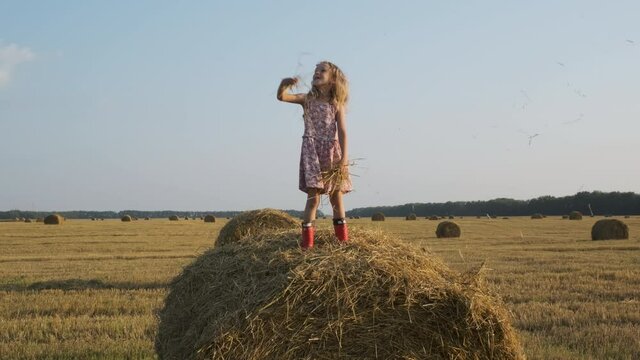 Cute 7-years Old Little Girl Throwing Up Straw while Standing on a Hay Bale in a Field, Harvest Time. Happy Childhood and Freedom Concept